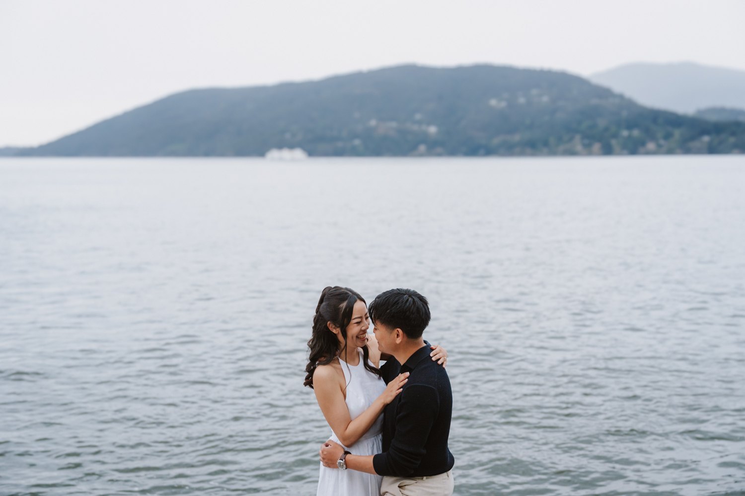 Aneeta and Christian in a quiet embrace at the water's edge, an island of Howe Sound behind them