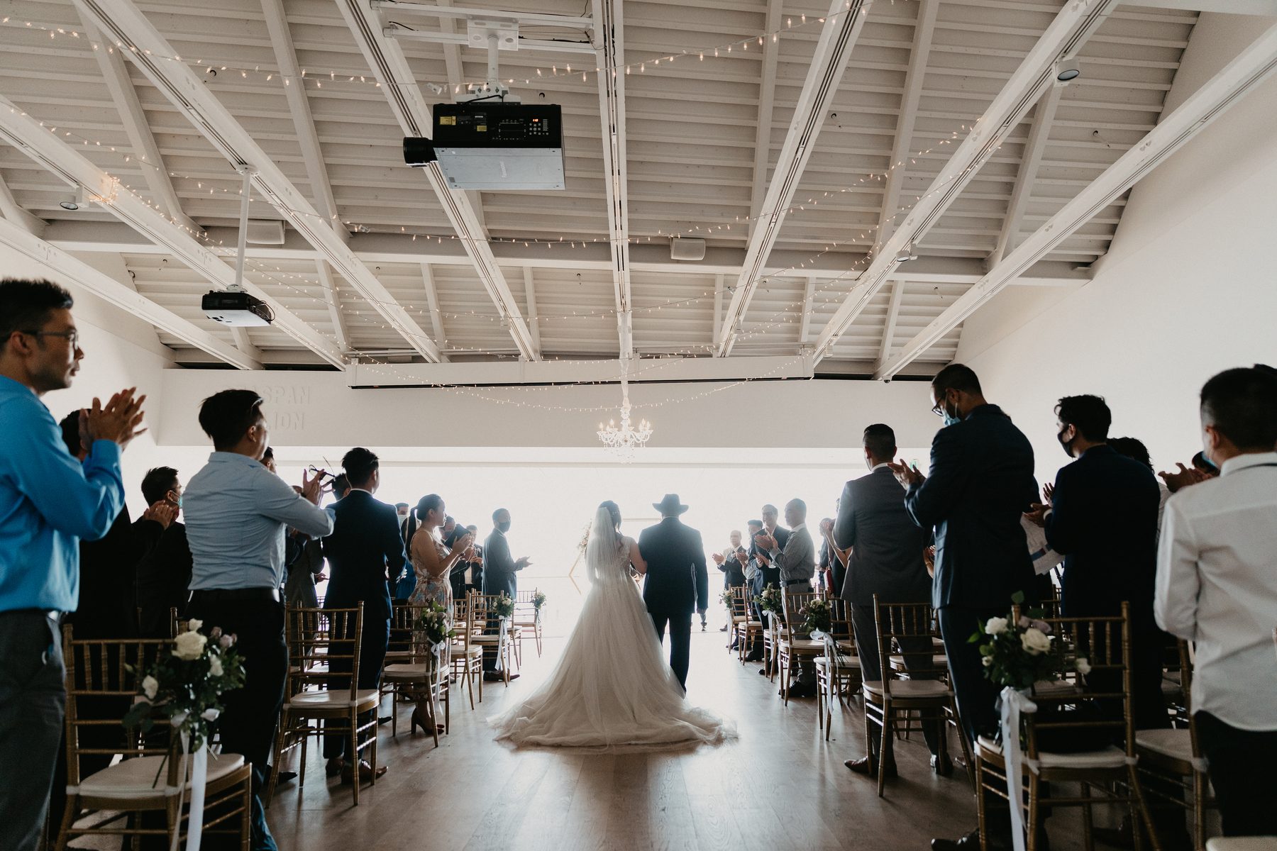 Bride walking down the aisle in a bright white room while guests stand and applaud