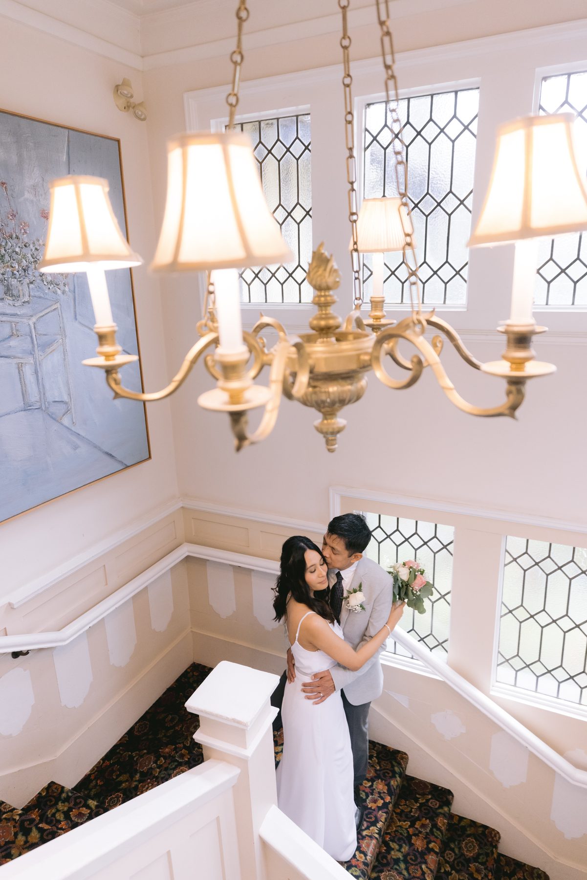 Couple holding each other on a landing beneath a chandelier, seen from the stairs above