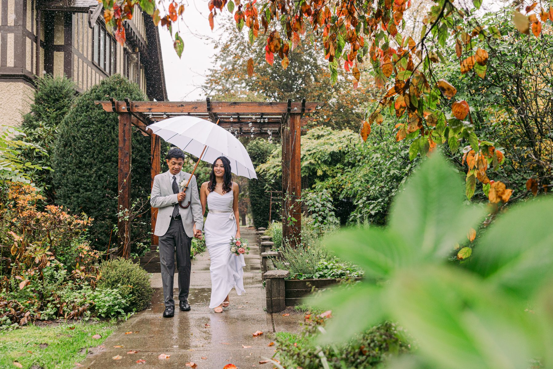 Bride and groom on a wet garden path sharing one white umbrella beneath autumn leaves