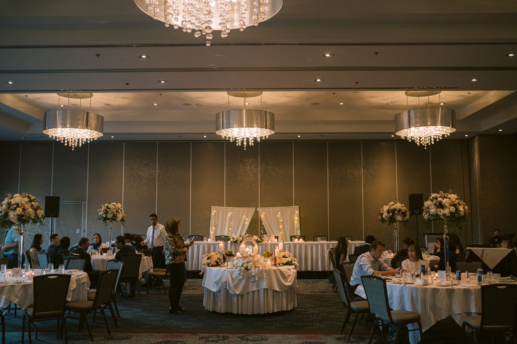 Ballroom set with round tables and low centrepieces under chandeliers as guests take their seats