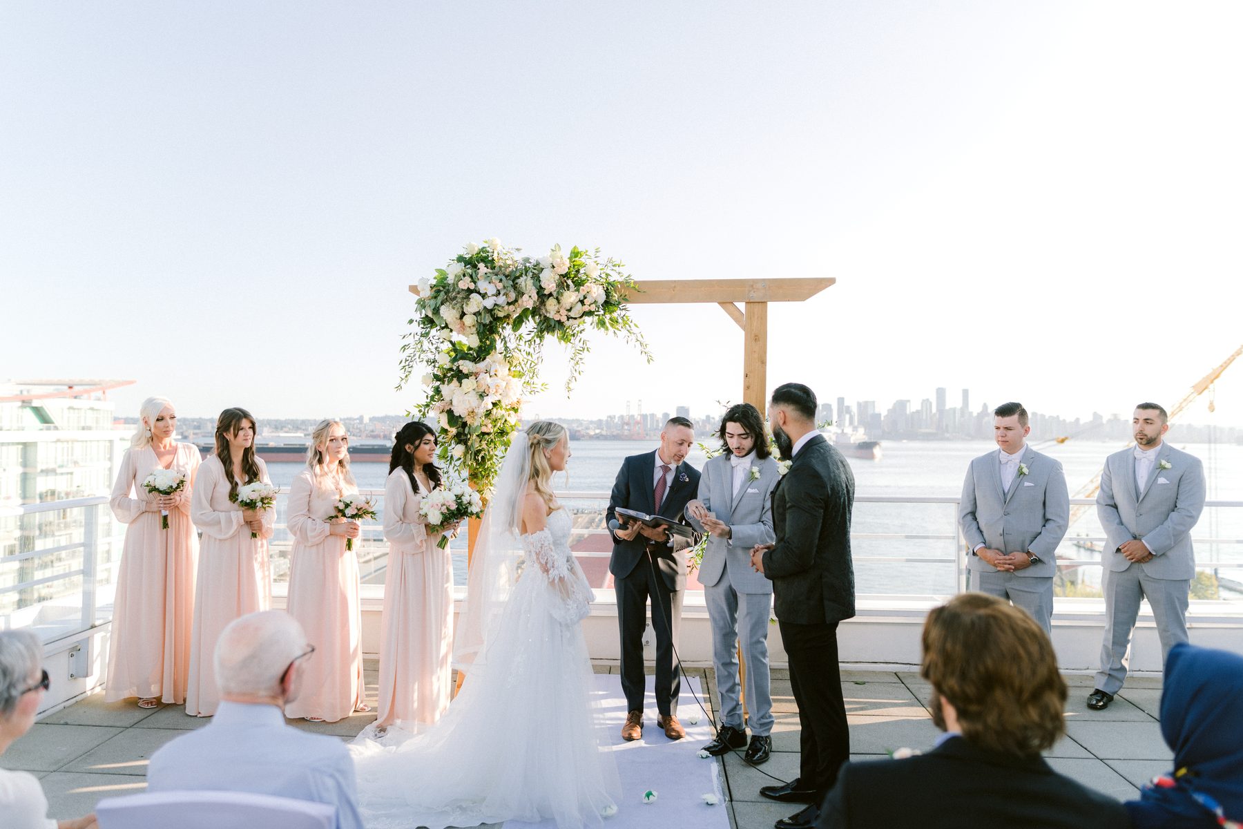 Wide view of a rooftop ceremony in progress with guests seated and the inlet stretching out behind the couple