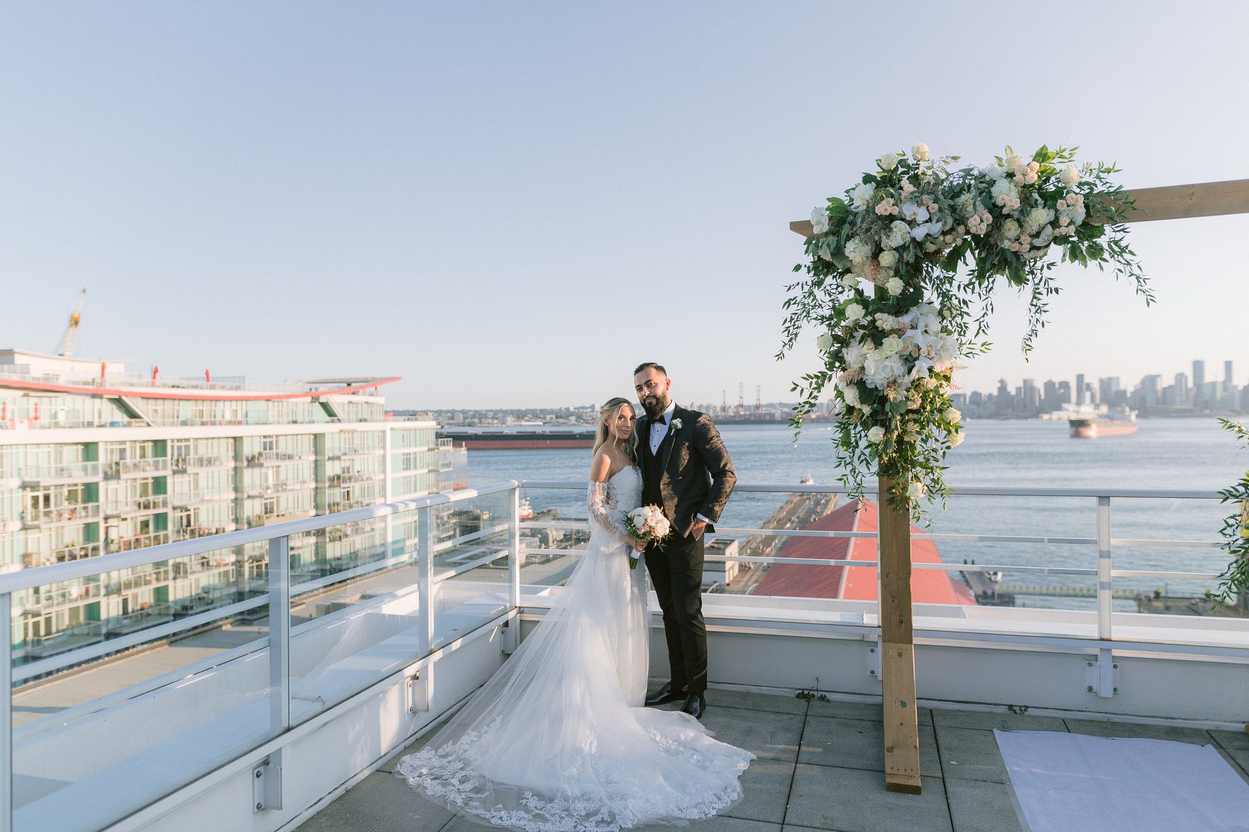 Bride and groom standing on an open rooftop deck with Burrard Inlet and the downtown Vancouver skyline behind them