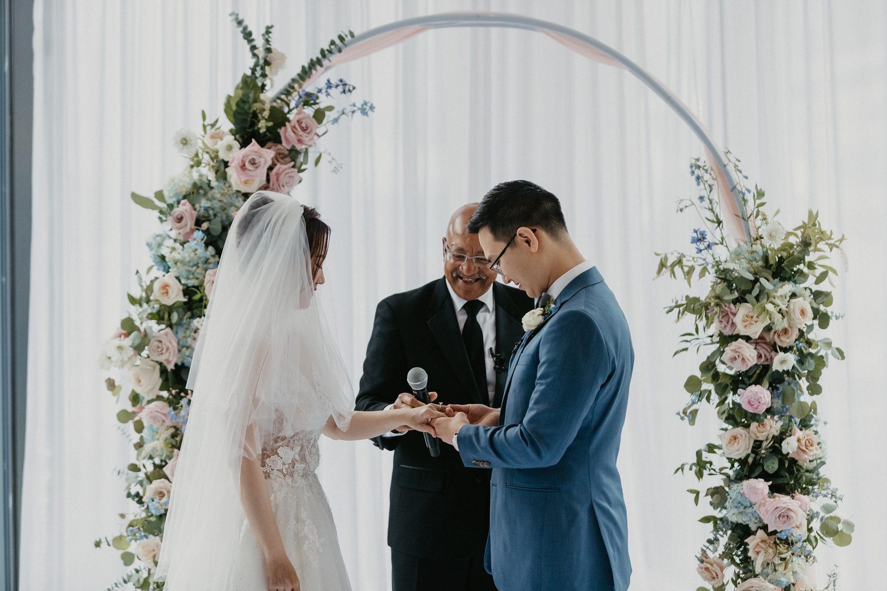 Groom sliding a ring onto his bride's finger beneath a curved floral arch