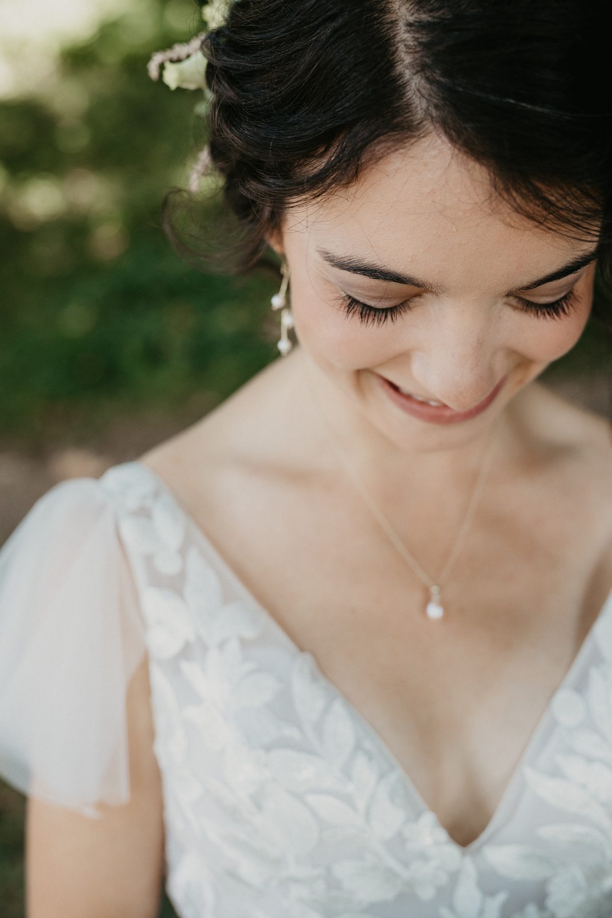 Bride in a lace gown looking down at her bouquet in dappled shade under summer trees