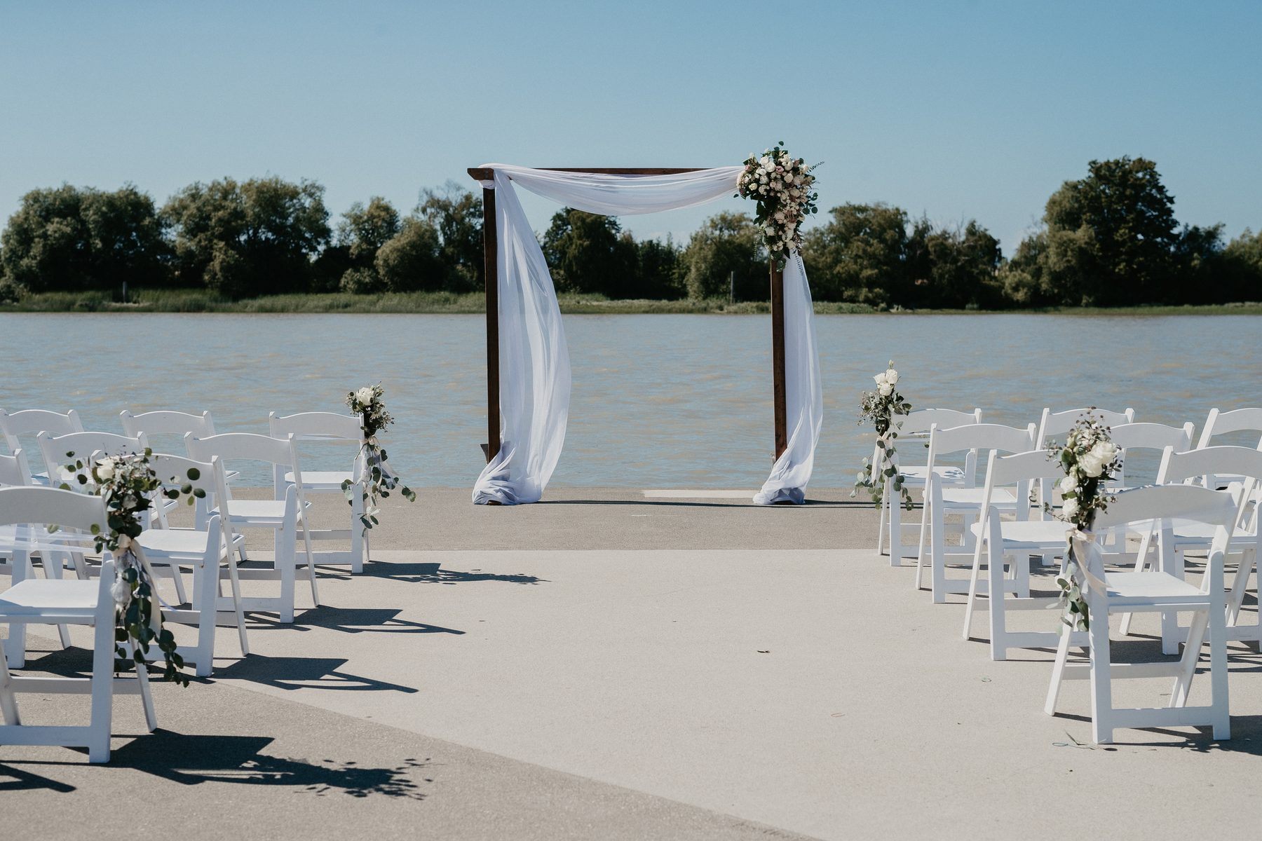 A simple wooden ceremony arch with white florals set up on a waterfront dock with rows of white chairs