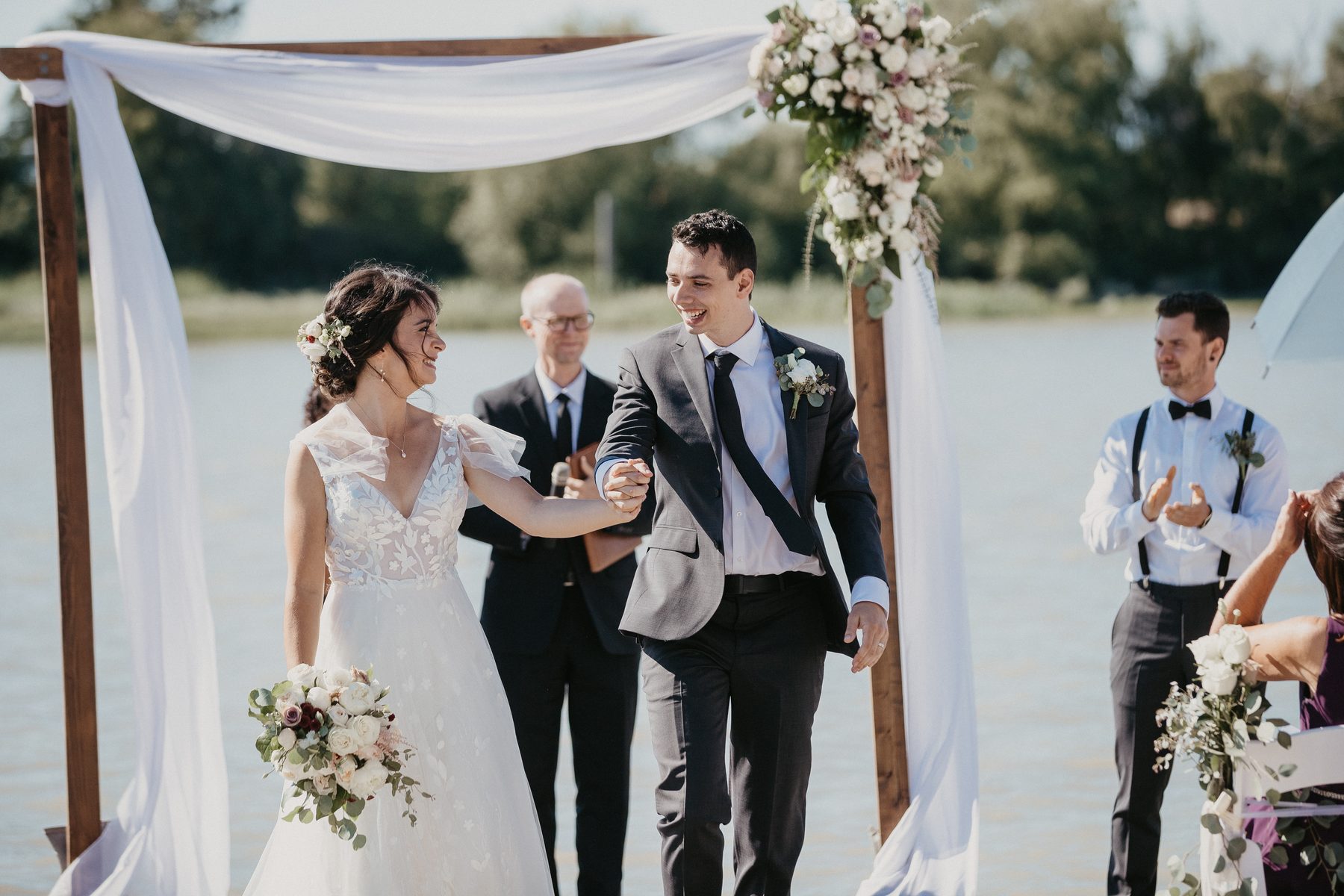 Couple facing each other during their vows on the waterfront while seated guests shelter under umbrellas