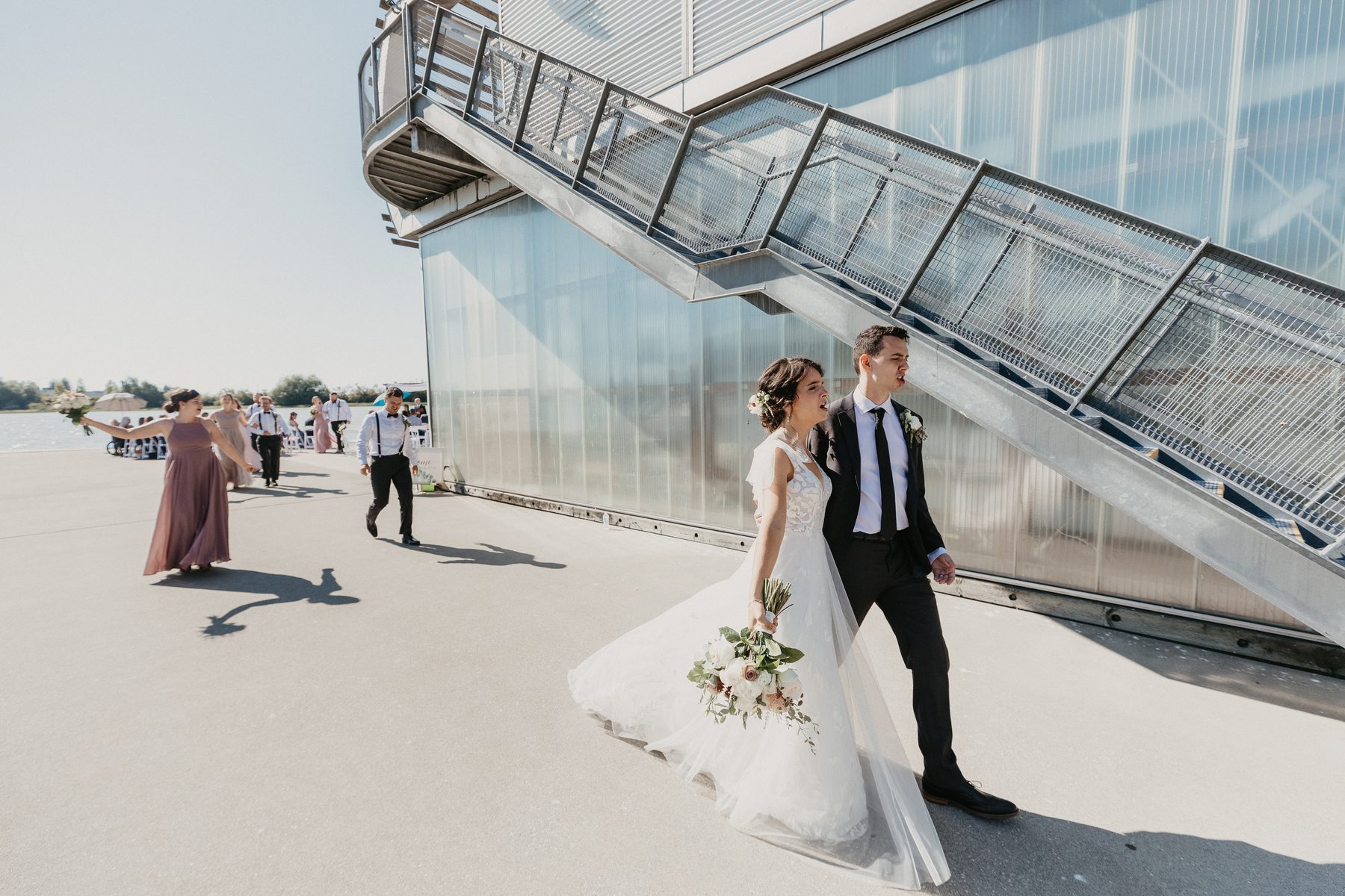 Bride and groom walking across a concrete dock past the glass wall of a riverside boathouse, wedding party following behind