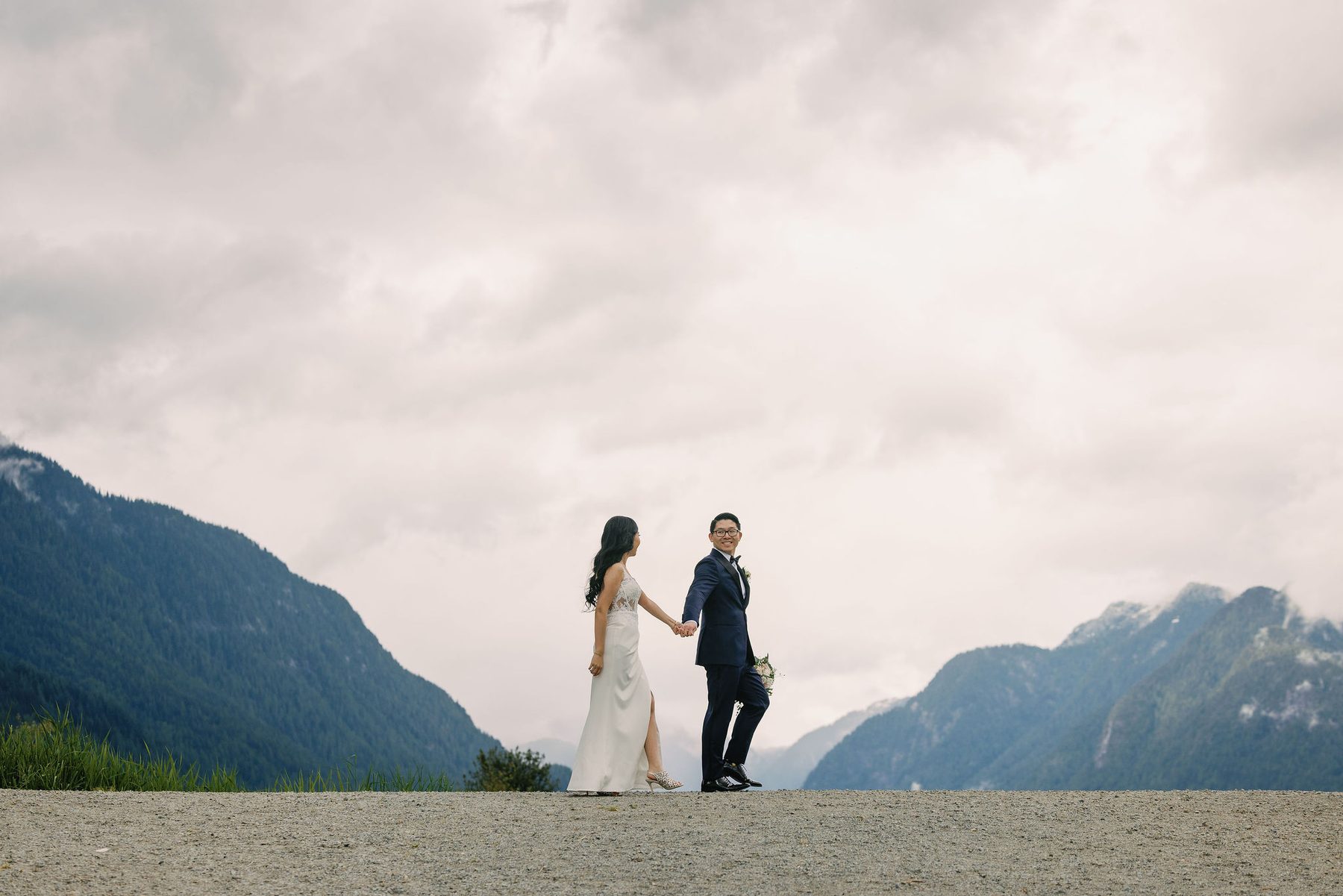 Bride and groom standing small against a wide backdrop of BC mountains and cloud