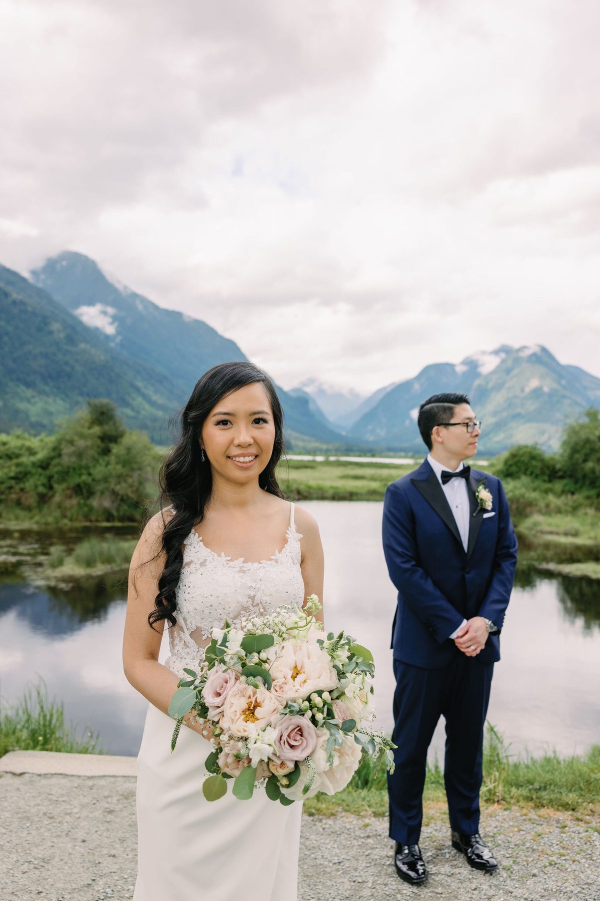 Bride and groom beside still water with the Coast Mountains reflected in it